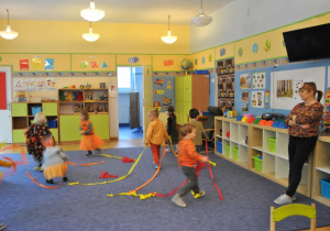 Dzieci tańczą w kole trzymając długie wstążki z kolorowej bibuły. Z prawej strony stoi nauczycielka. Ujęcie 2